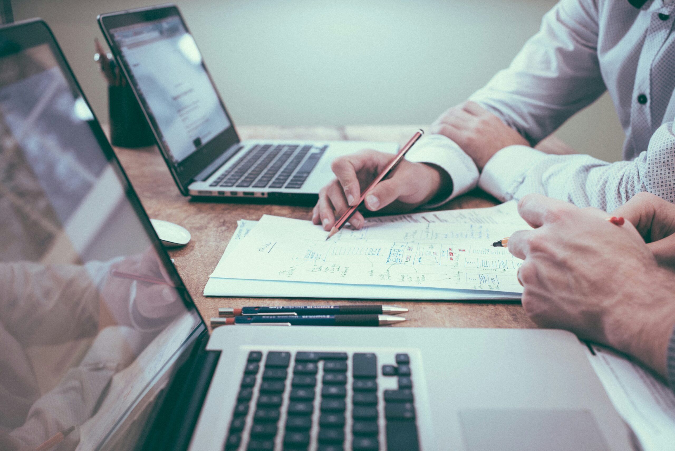 Two people collaborating over documents with laptops on a wooden table.